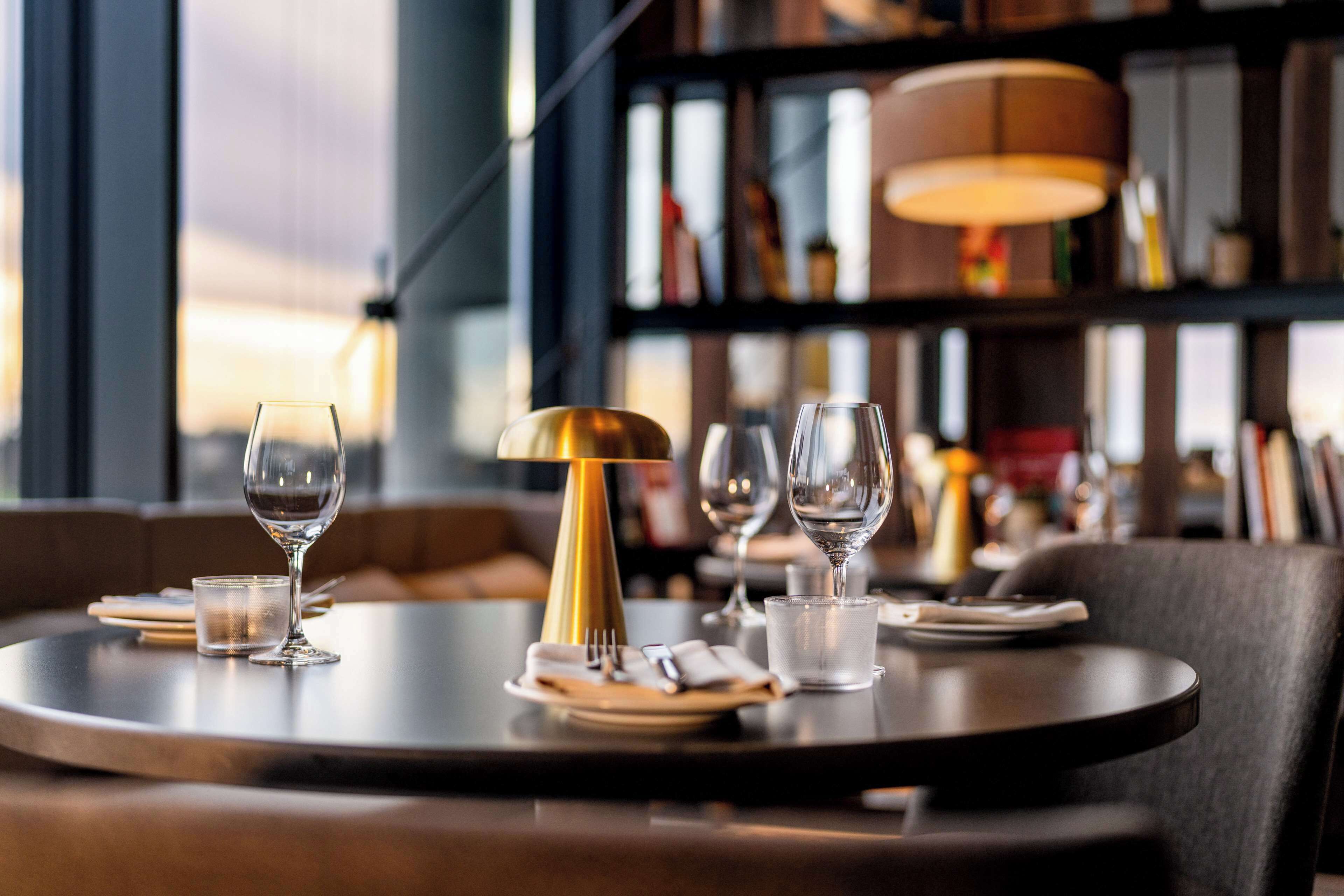 A dining table set with wine glasses, a water glass, and cutlery, with a bookshelf and a lamp in the background.