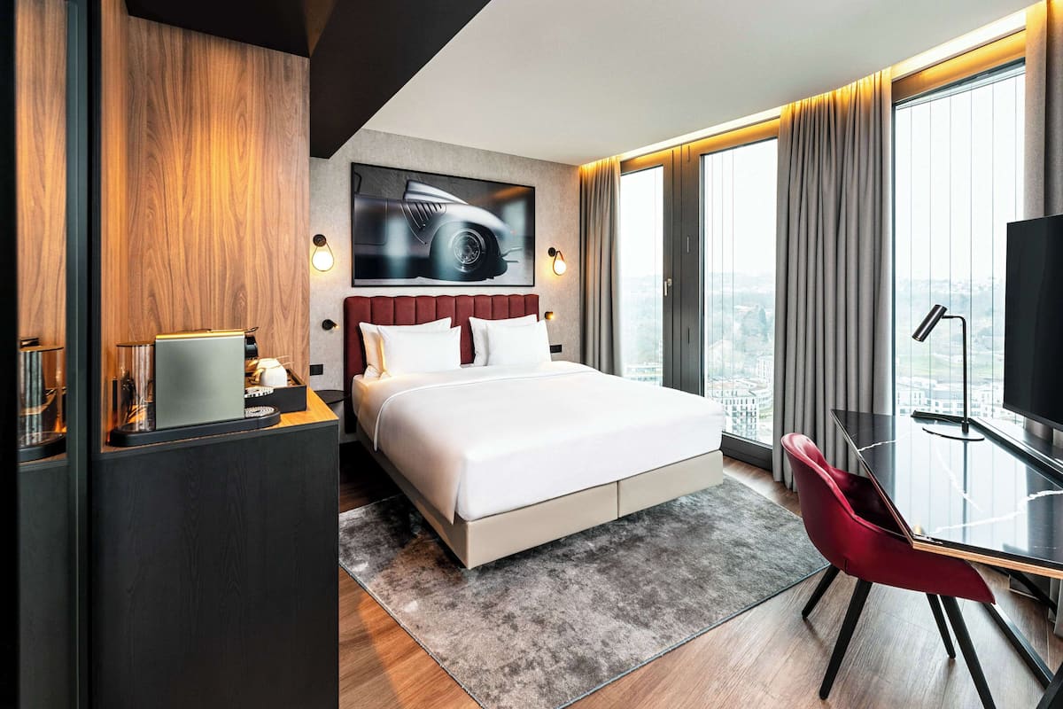 A modern hotel room with a large bed, a desk with a chair, a television, and a city view.