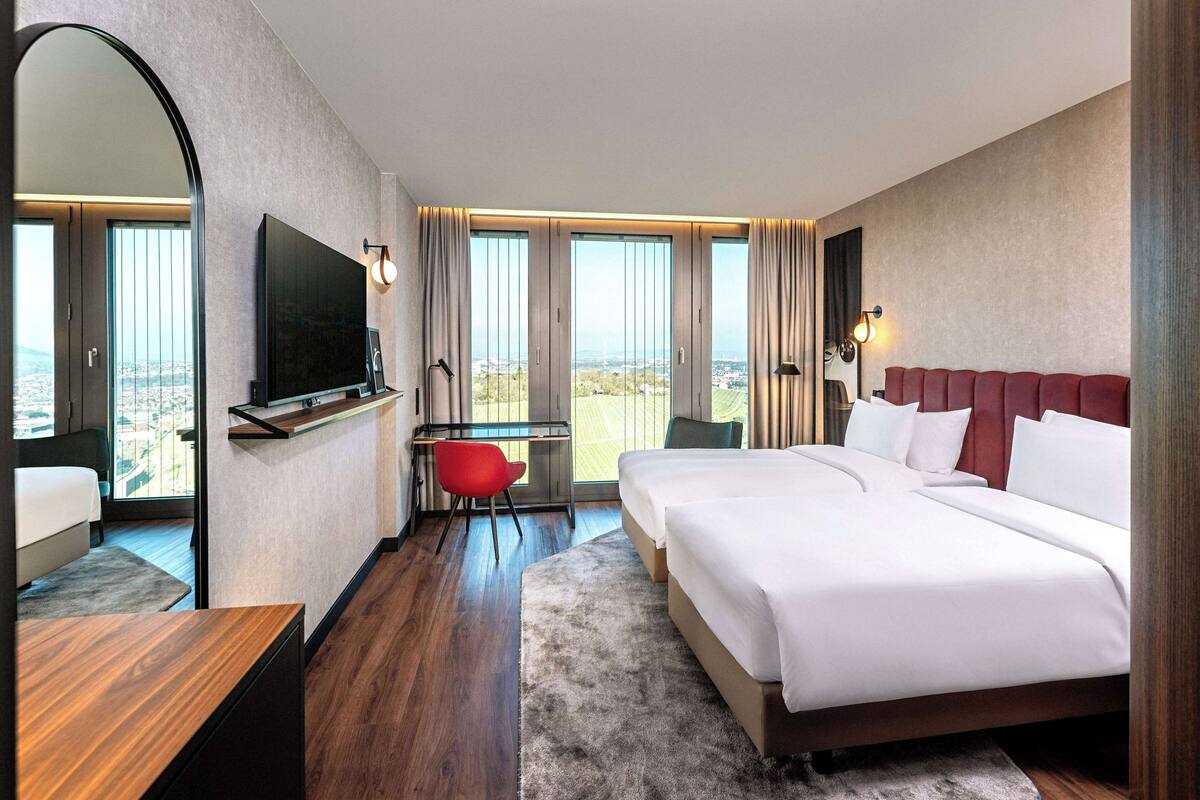 A hotel room with a large bed, a desk with a chair, a TV mounted on the wall, and a view of the city through large windows.