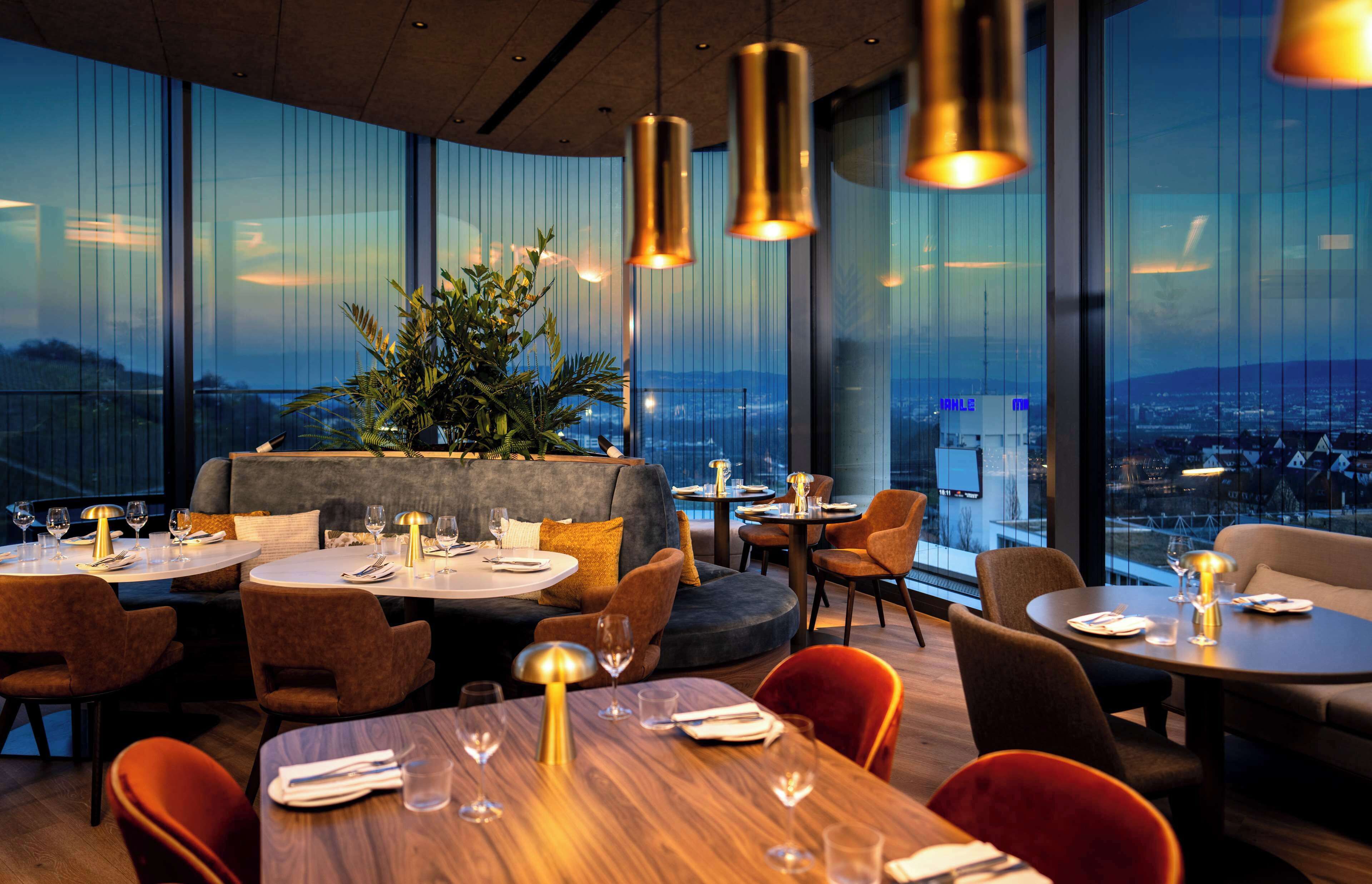 Ein Restaurant mit runden Tischen und Stühlen, einer großen Pflanzenarrangement in der Mitte und Blick auf die Stadt durch große Fenster.