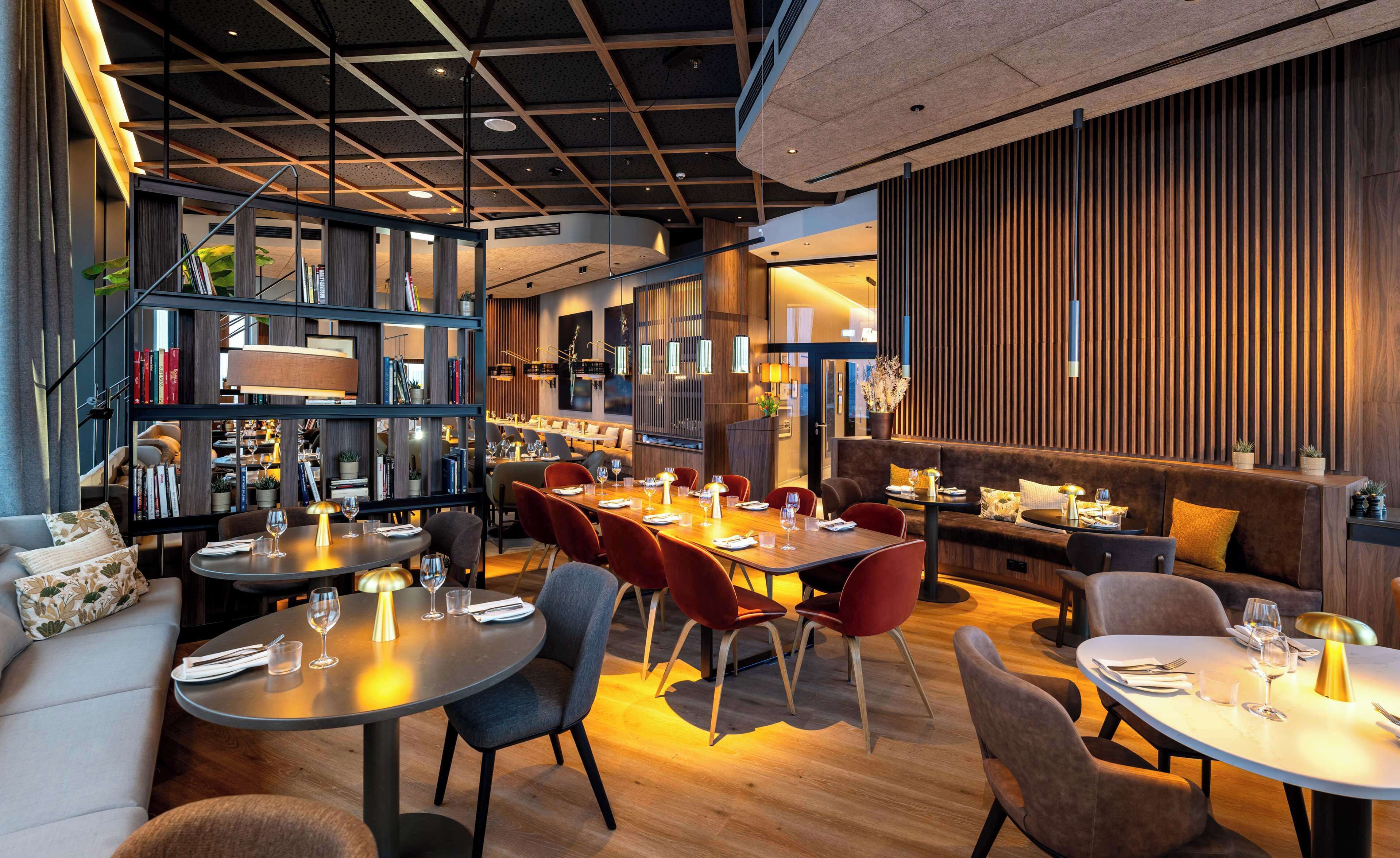 Un restaurante moderno con diversas opciones de asientos, incluyendo mesas redondas y un área con sofás. Se observa una estantería llena de libros y objetos decorativos.