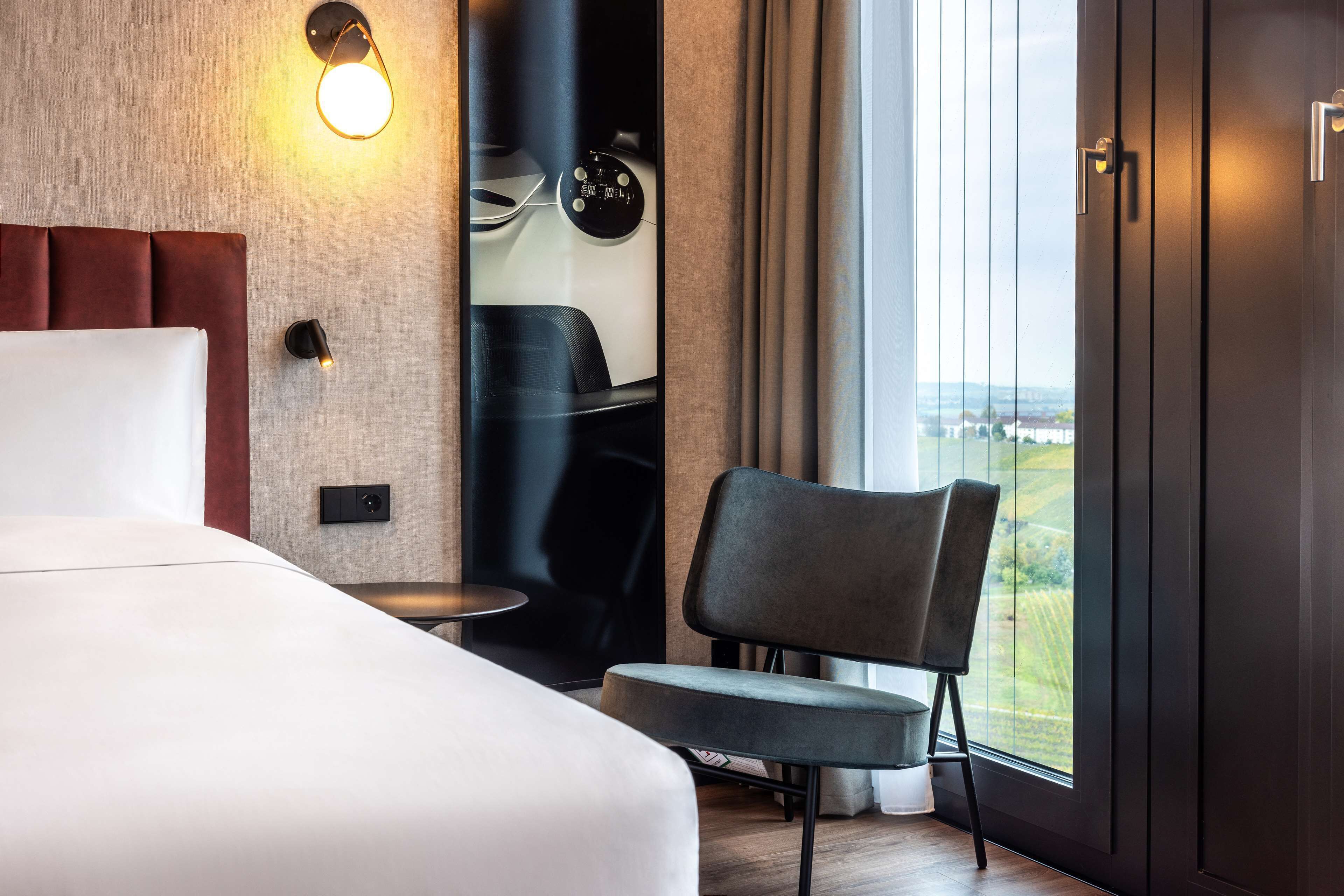 A modern hotel room with a large bed, a chair, a small table, and a view of the countryside through a large window.