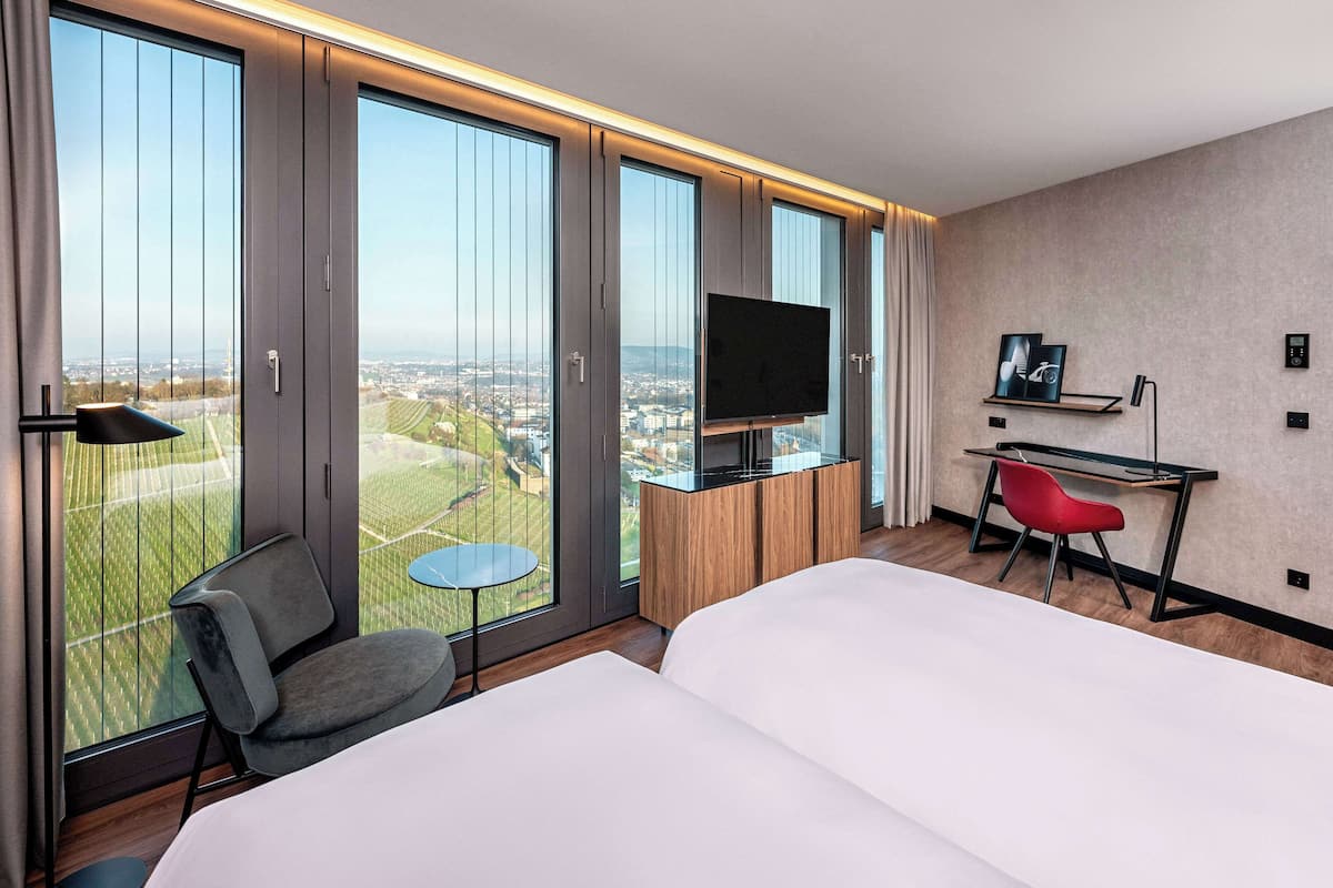 A hotel room with a large bed, a desk with a chair, a television, and a view of the city through large windows.