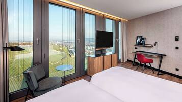 A hotel room with a large bed, a desk with a chair, a television, and a view of the city through large windows.