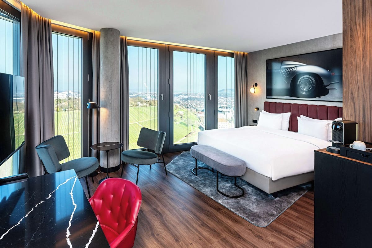 A modern hotel room with a large bed, a seating area with chairs, a dining table, and a city view through large windows.