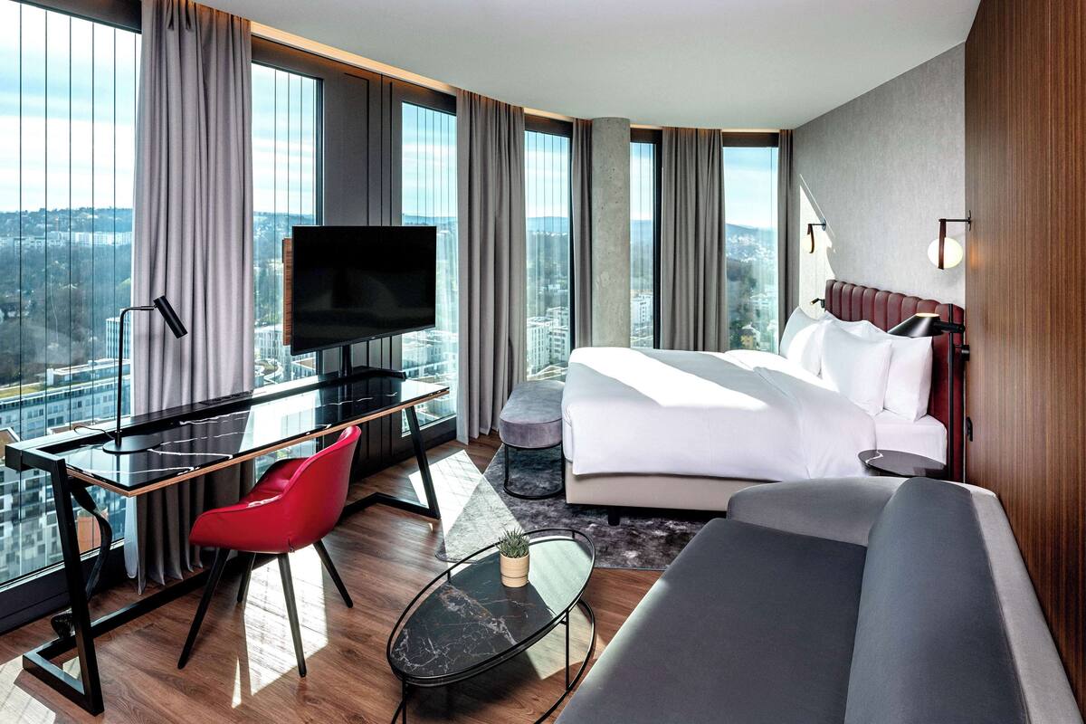A modern hotel room with a large bed, a desk with a TV, a red chair, and a view of the city.
