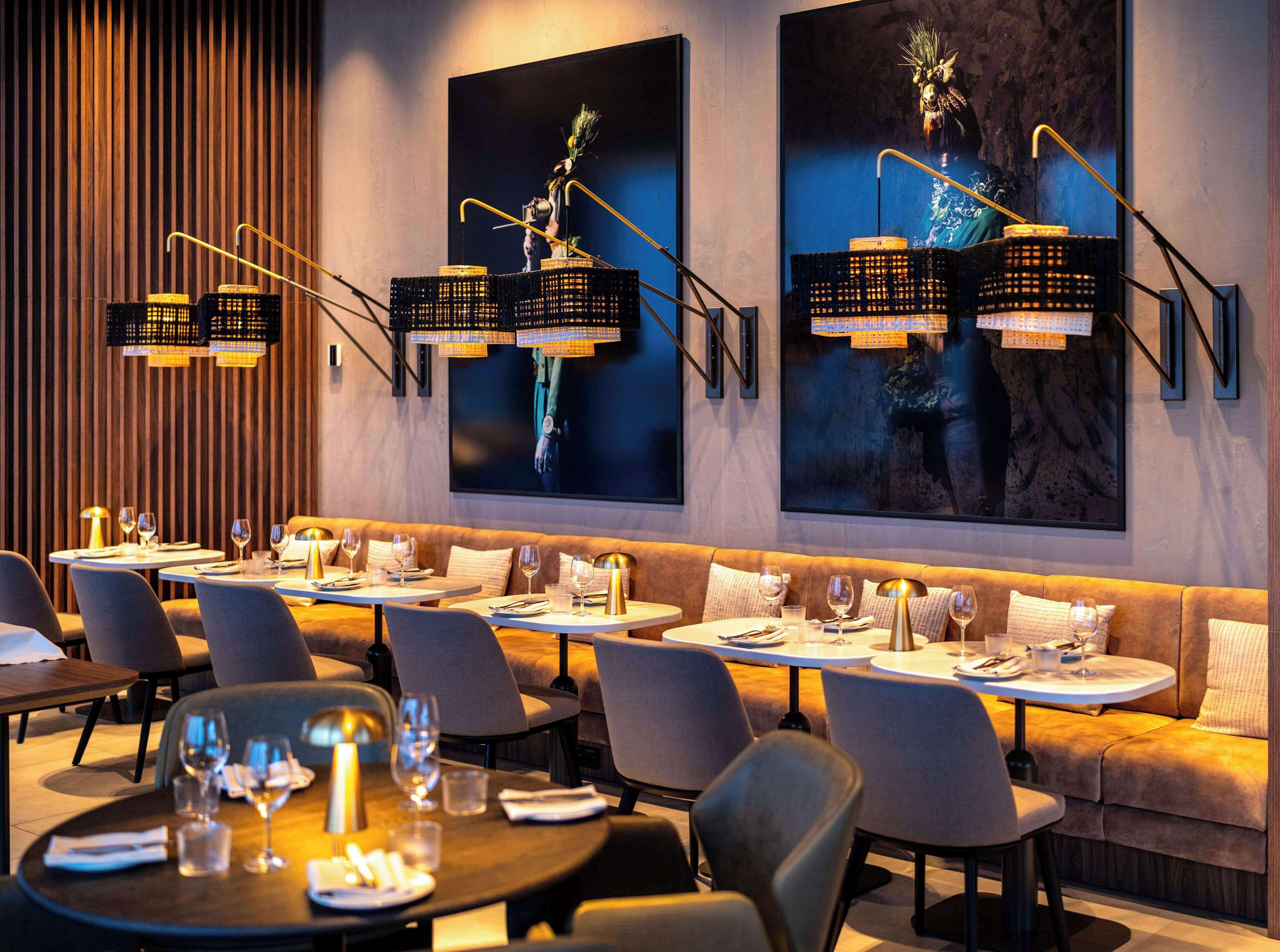 Un restaurante moderno con mesas altas y asientos acolchados, que cuenta con luminarias contemporáneas y grandes obras de arte en la pared.