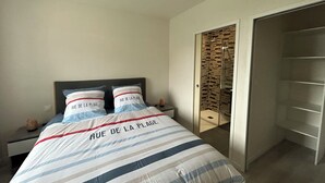 Ett sovrum med en säng som har ett randigt överkast och kuddar med texten "RUE DE LA PLAGE", en glasbeklädd walk-in-garderob och en dörr som leder till ett badrum med dusch.