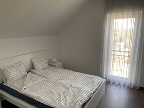 Habitació individual amb un llit i capçalera senzilla, una taula de nit amb una làmpada i una finestra amb cortines de tul.
