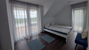 Een slaapkamer met een bed, twee ramen met dunne gordijnen, een balkon en een klein tafeltje met een stoel.