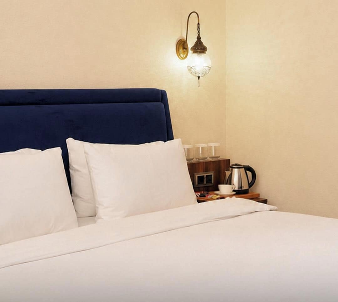 A hotel room with a bed, pillows, a nightstand, a kettle, and a wall-mounted light fixture.