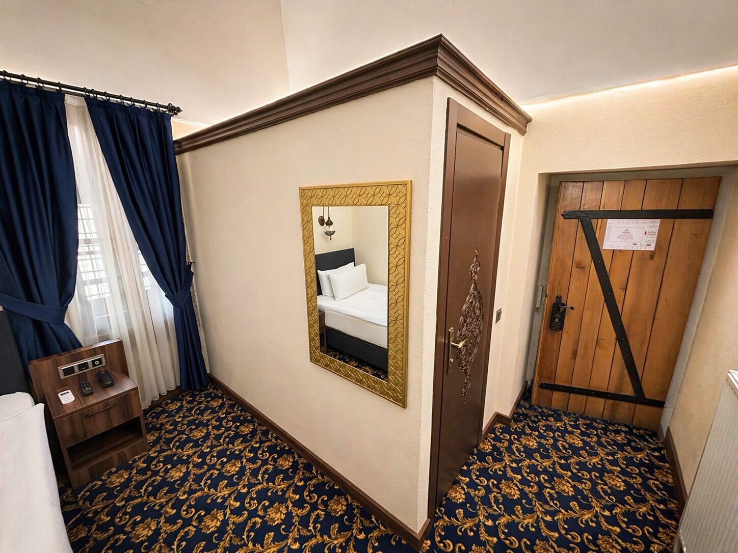 Camera d'albergo con un letto, uno specchio, una porta in legno, una scrivania con un telefono e una moquette fantasia.