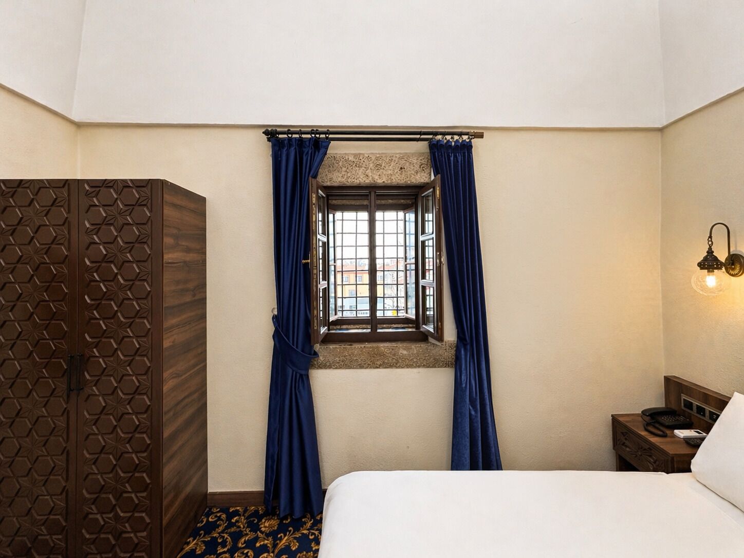 Una habitación de hotel con cama, ventana con cortinas azules, armario de madera, mesita de noche con lámpara, y una lámpara de pared.