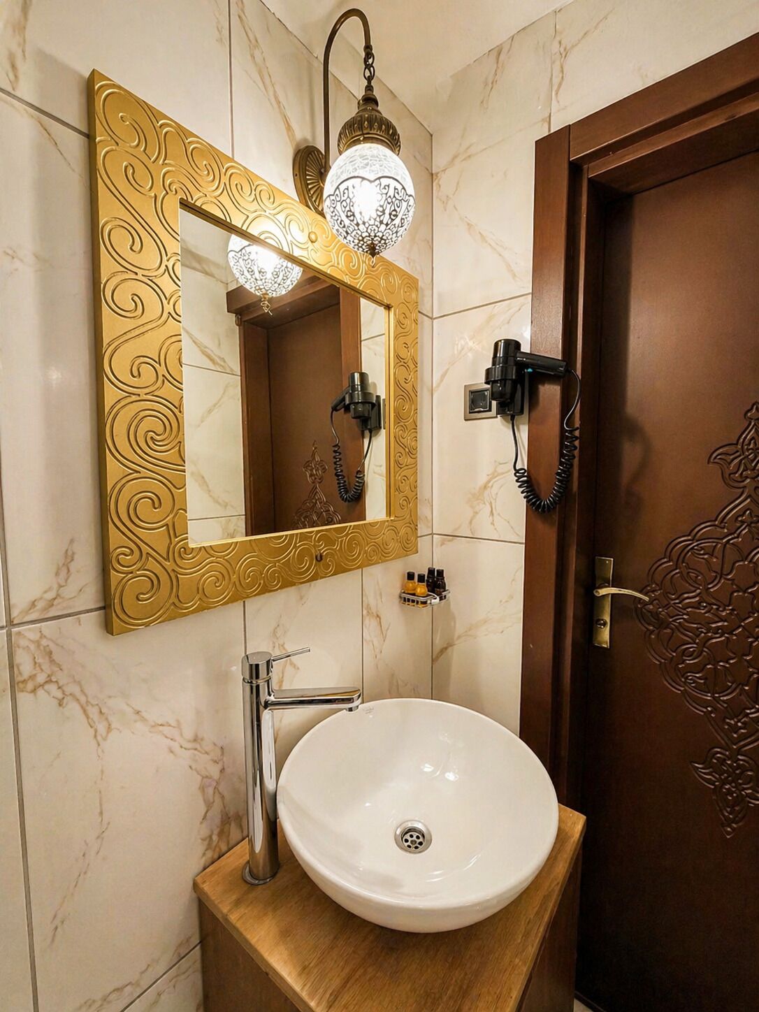 Casa de banho com parede de mármore, espelho com moldura dourada, lavatório branco e porta com detalhes entalhados.