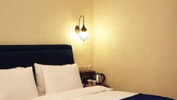 Een net opgemaakt bed met witte en marineblauwe beddengoed, een wandlamp en een klein tafeltje met een koffiezetapparaat en een kop.