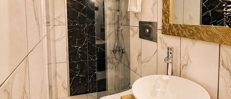 A modern bathroom with a marble floor, a white sink, a glass shower enclosure, and a large mirror with a gold frame.