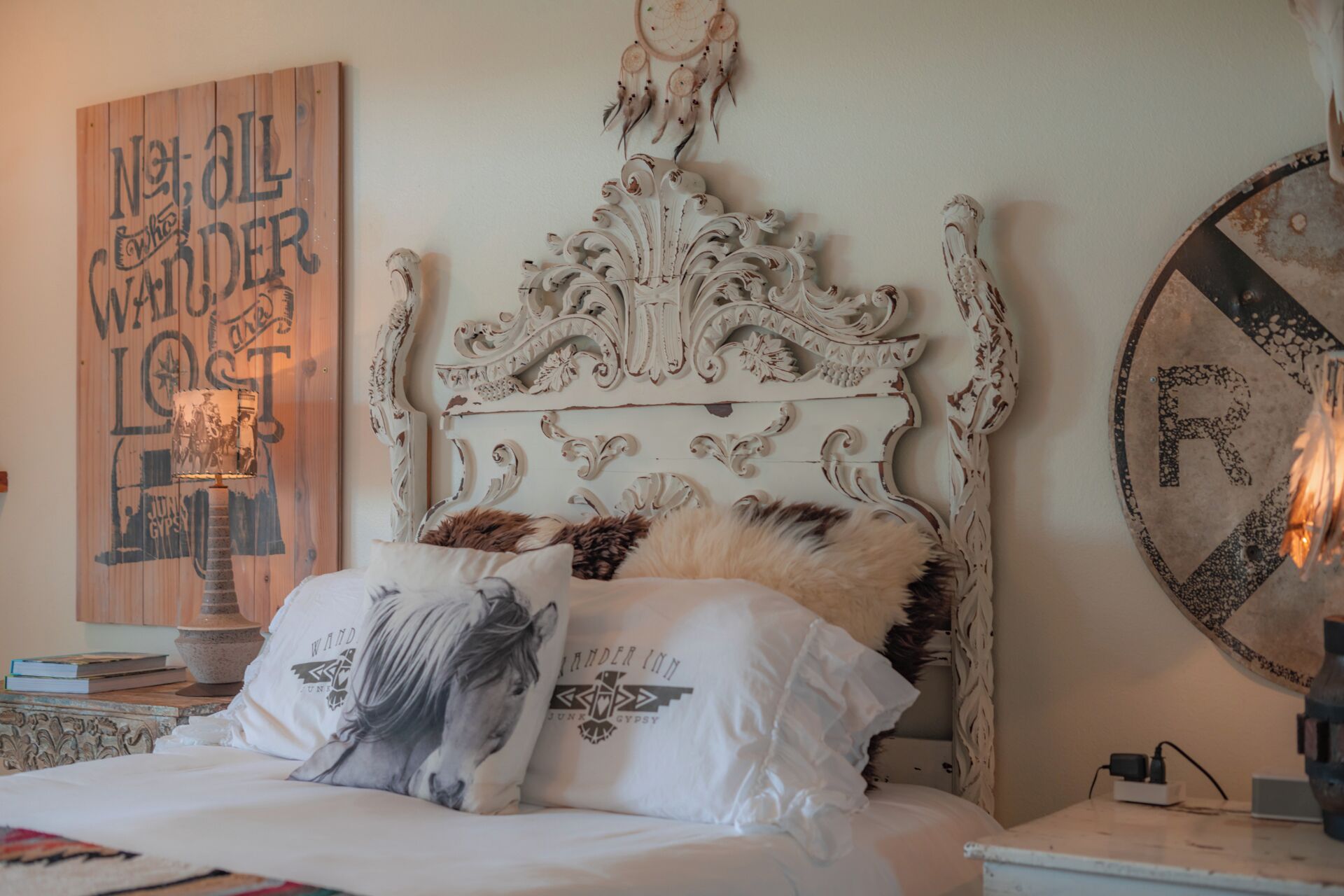A bedroom with an ornate white headboard, a bed with white bedding and a horse-themed pillow, a wall with a dreamcatcher, and a round mirror on the nightstand.