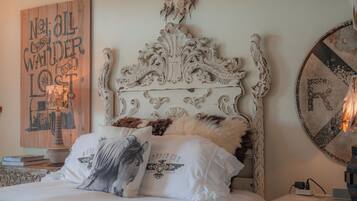 A bedroom with an ornate white headboard, a bed with white bedding and a horse-themed pillow, a wall with a dreamcatcher, and a round mirror on the nightstand.