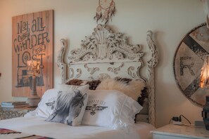 A bedroom with an ornate white headboard, a bed with white bedding and a horse-themed pillow, a wall with a dreamcatcher, and a round mirror on the nightstand. - The Wanderlust Room - Wander Inn (Round Top)