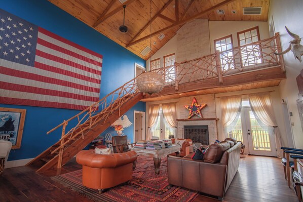 A spacious living room with a high ceiling, a large American flag, a wooden staircase, a fireplace, and a star decoration. - The Bandita Room - Wander Inn (Round Top)