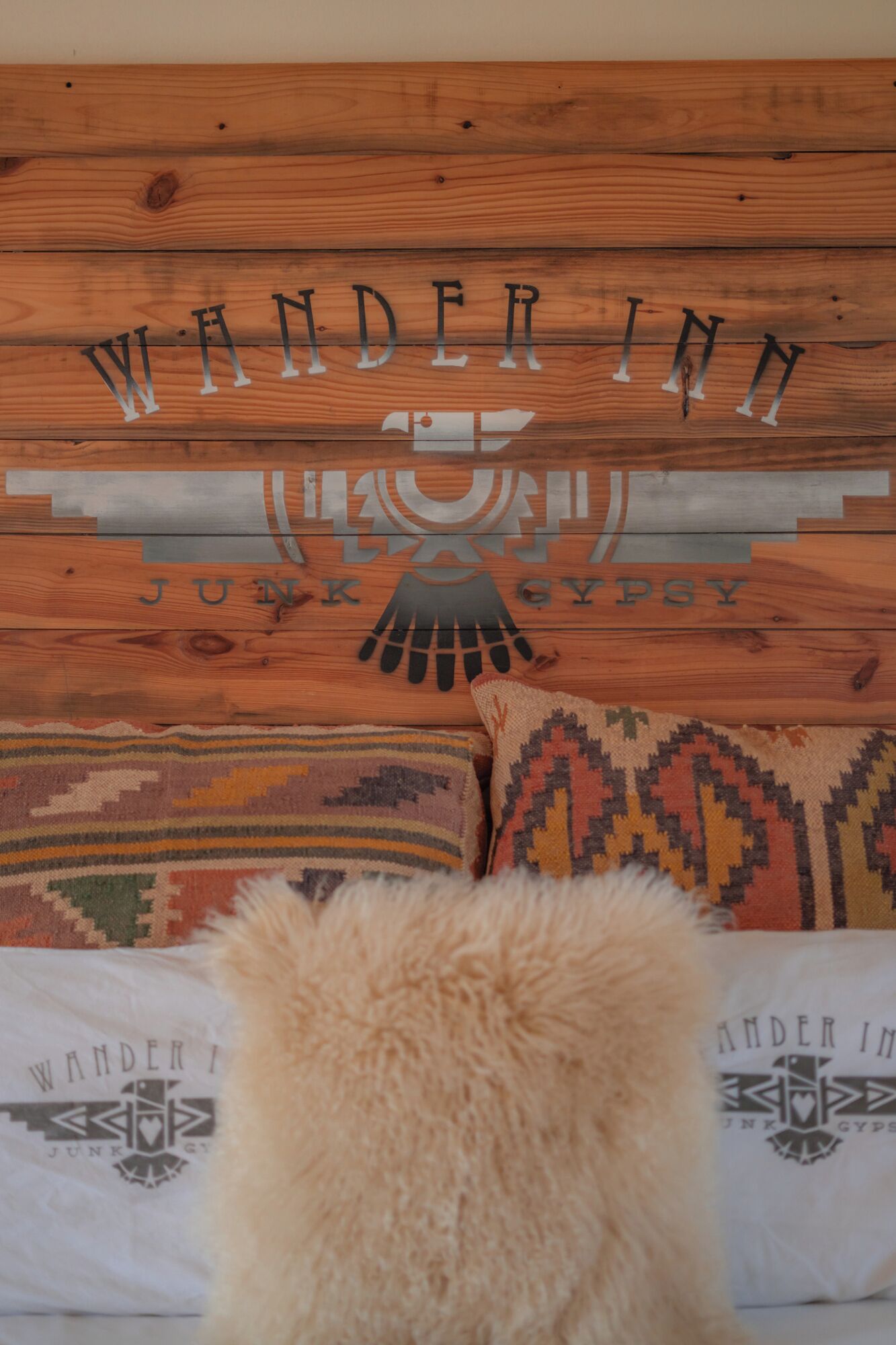 A bed with a wooden headboard displaying the text "WANDER IN JUNK GYPSY" and a fluffy pillow in front.