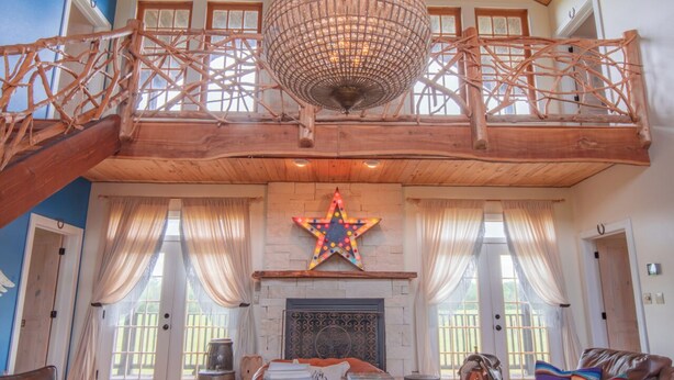A spacious living room with a high ceiling, a large chandelier, a fireplace, and a balcony with wooden railings. - The Stardust Room - Wander Inn (Round Top)