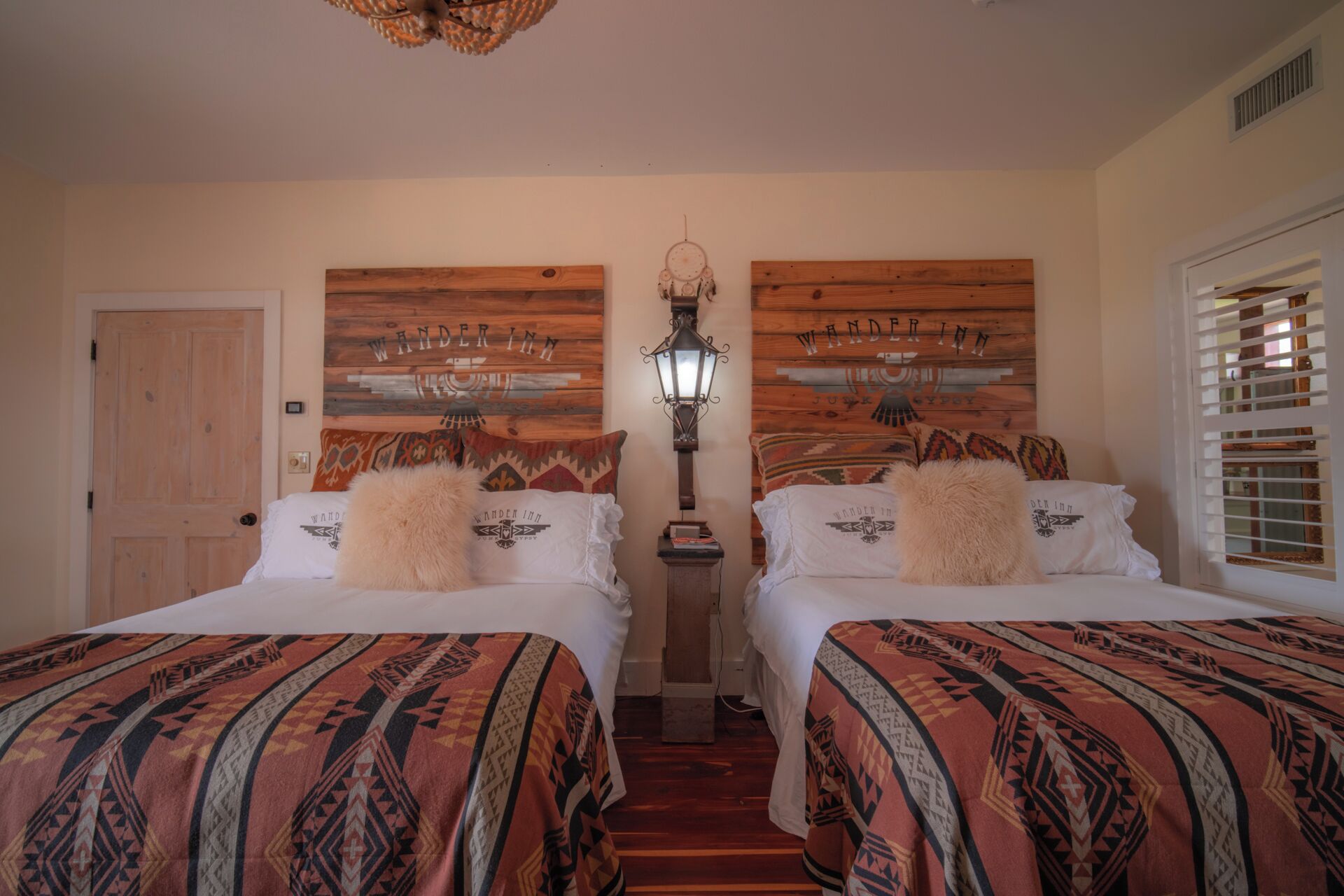 Una habitación con dos camas, cada una con ropa de cama y almohadas estampadas, un cabecero de madera con la marca 'Wander Inn', un farol colgando de la pared y una puerta.