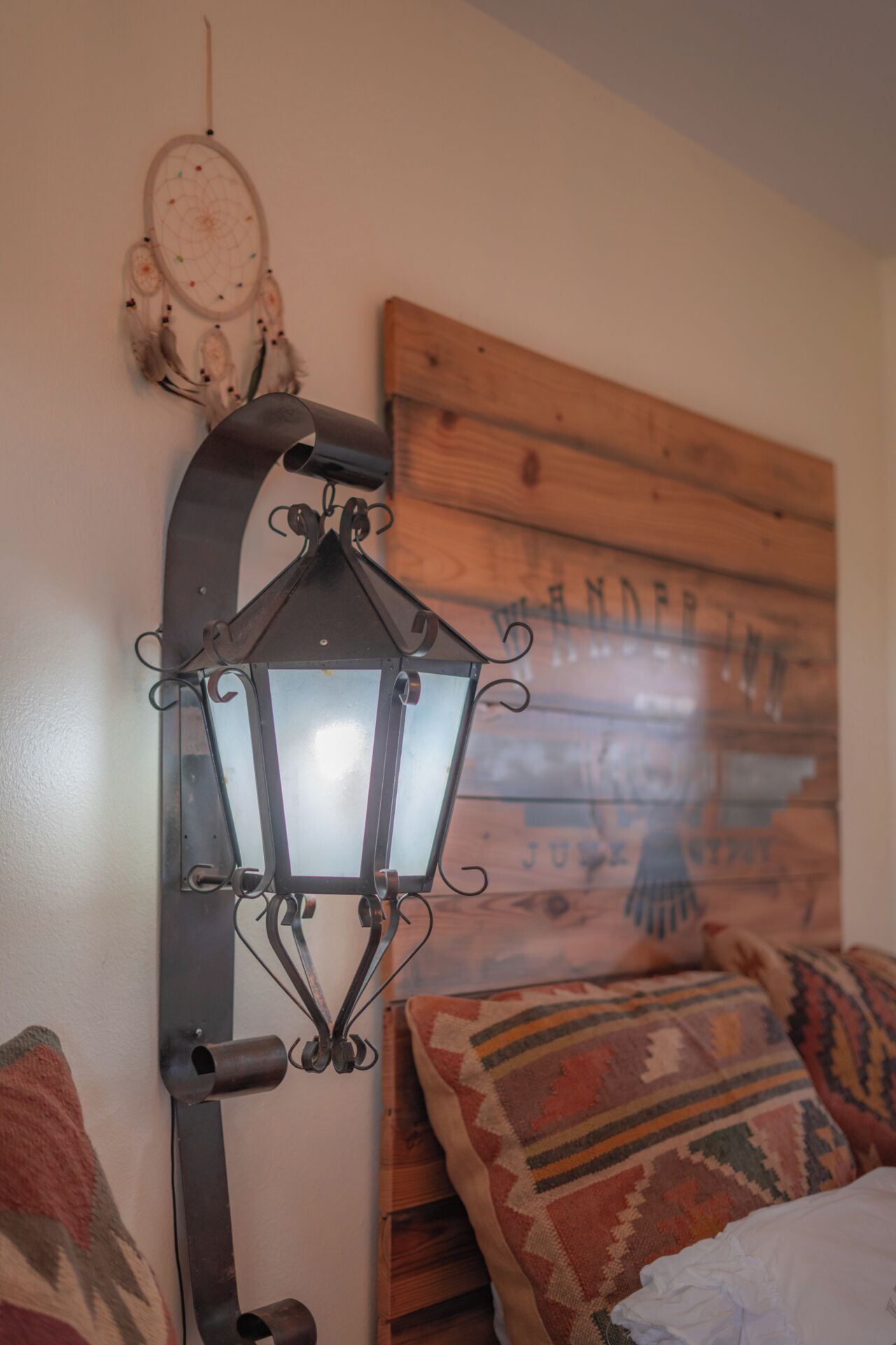 A room with a unique wall-mounted lantern, a wooden headboard with text, and patterned pillows.