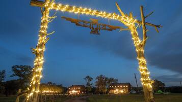 Une arche rustique en bois, décorée de guirlandes lumineuses, mène à un chemin de campagne bordé de maisons en arrière-plan.