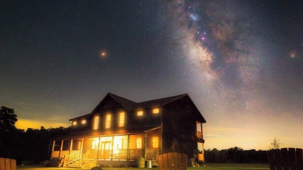 A large house with multiple windows and a well-lit porch, surrounded by a grassy yard and a fire pit. - The Rambler Room - Wander Inn (Round Top)