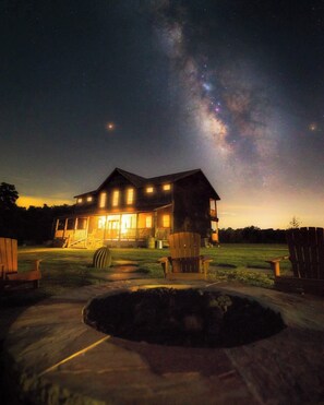 A large house with multiple windows and a well-lit porch, surrounded by a grassy yard and a fire pit.