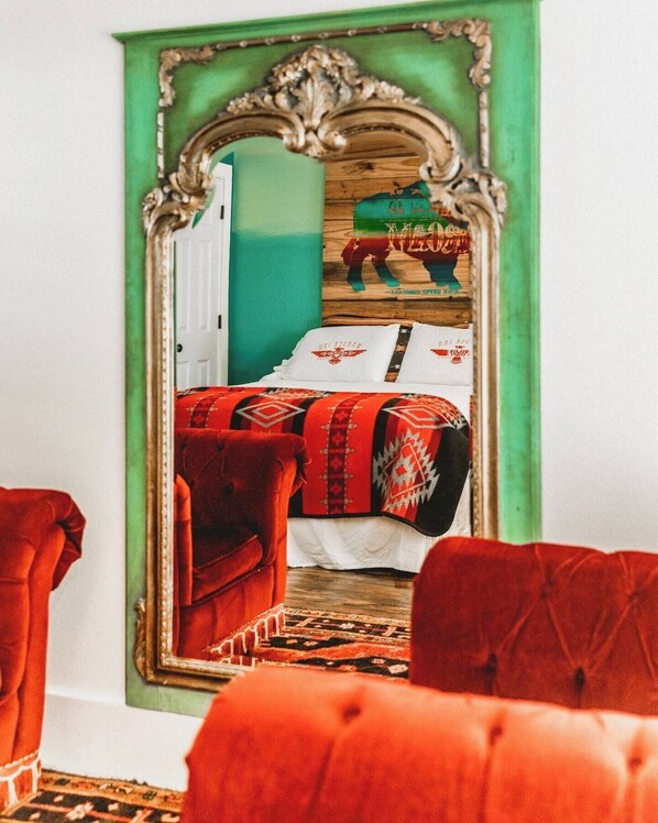A room with a large mirror reflecting a bed with patterned bedding, a red upholstered chair, and a wooden headboard. - The Vagabond Room - Wander Inn (Round Top)