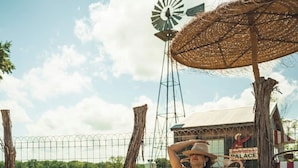 A person relaxing on a striped lounge chair under a woven umbrella, with a windmill and a building labeled "PALACE" in the background. - Gypsy Farmhouse - Wander Inn (Round Top)