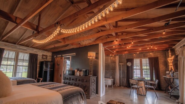 A rustic bedroom with a bed, a dresser, and a dining area with chairs and a table. - Thelma's Garage - Wander Inn (Round Top)