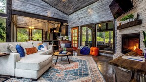 A spacious living room with a large sofa, a coffee table, and a fireplace. There are large windows and a stone wall. - 2Bdr with elevated design and bunkhouse! (Broken Bow)
