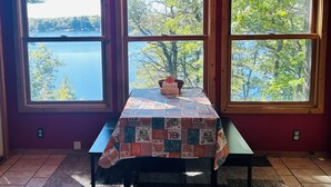 Een kamer met drie grote ramen met uitzicht op een meer en bomen, een tafel met een quilt en een plafondventilator.