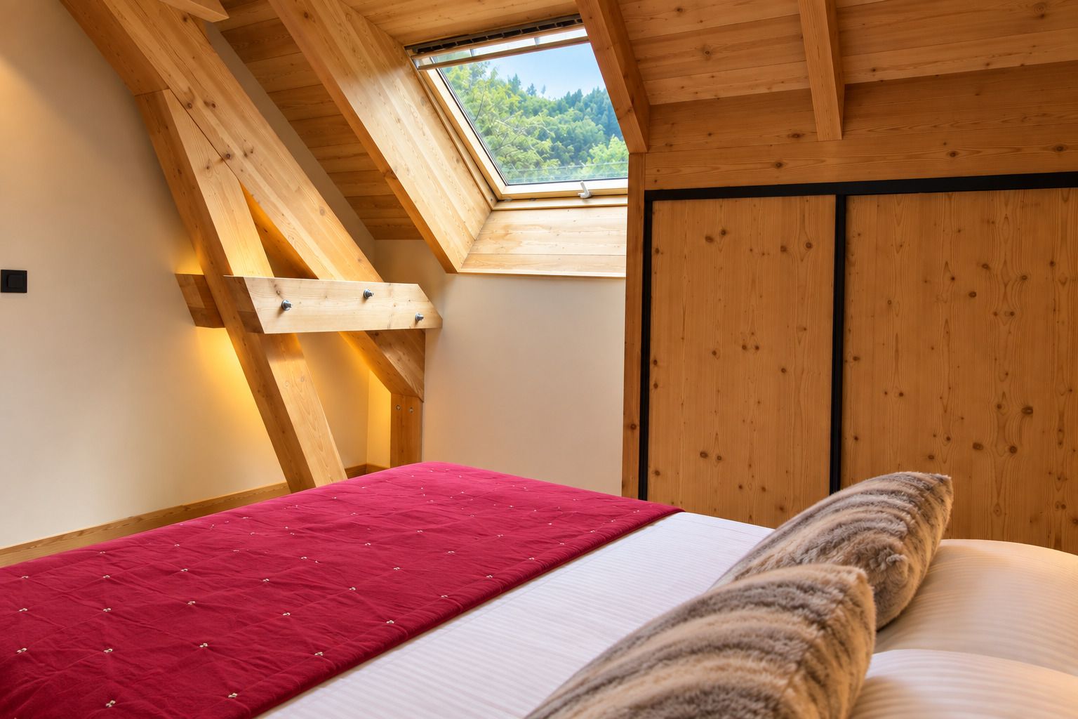 A bedroom with a sloped ceiling, a skylight, a bed with a red bedspread, and wooden walls.