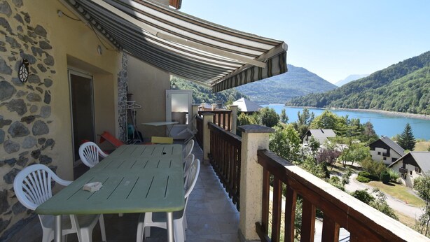 A balcony with a view of a lake and mountains, a table with chairs, and a stone wall. - Chalet Roches Grandes 47 (Vaujany)