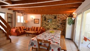 A cozy living area with a stone wall, wooden beams, a dining table with patterned tablecloth, and a staircase leading upstairs. - Chalet Roches Grandes 47 (Vaujany)