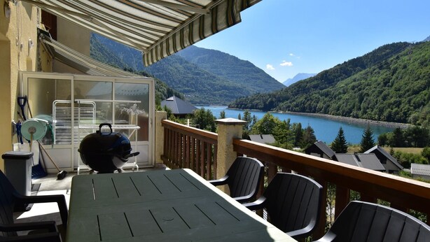 A balcony with a table and chairs overlooking a lake and mountains. - Chalet Roches Grandes 49 (Vaujany)