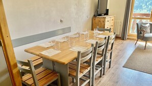 A dining area with a wooden table and chairs, a television on a wooden cabinet, and a view of greenery outside. - Appartement de La Fare, Apt 30 (Vaujany)
