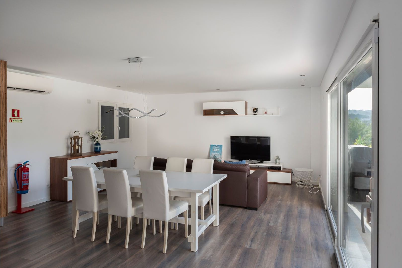 A modern living room with a dining area, a flat-screen TV, and a large window offering a view of the outdoors.