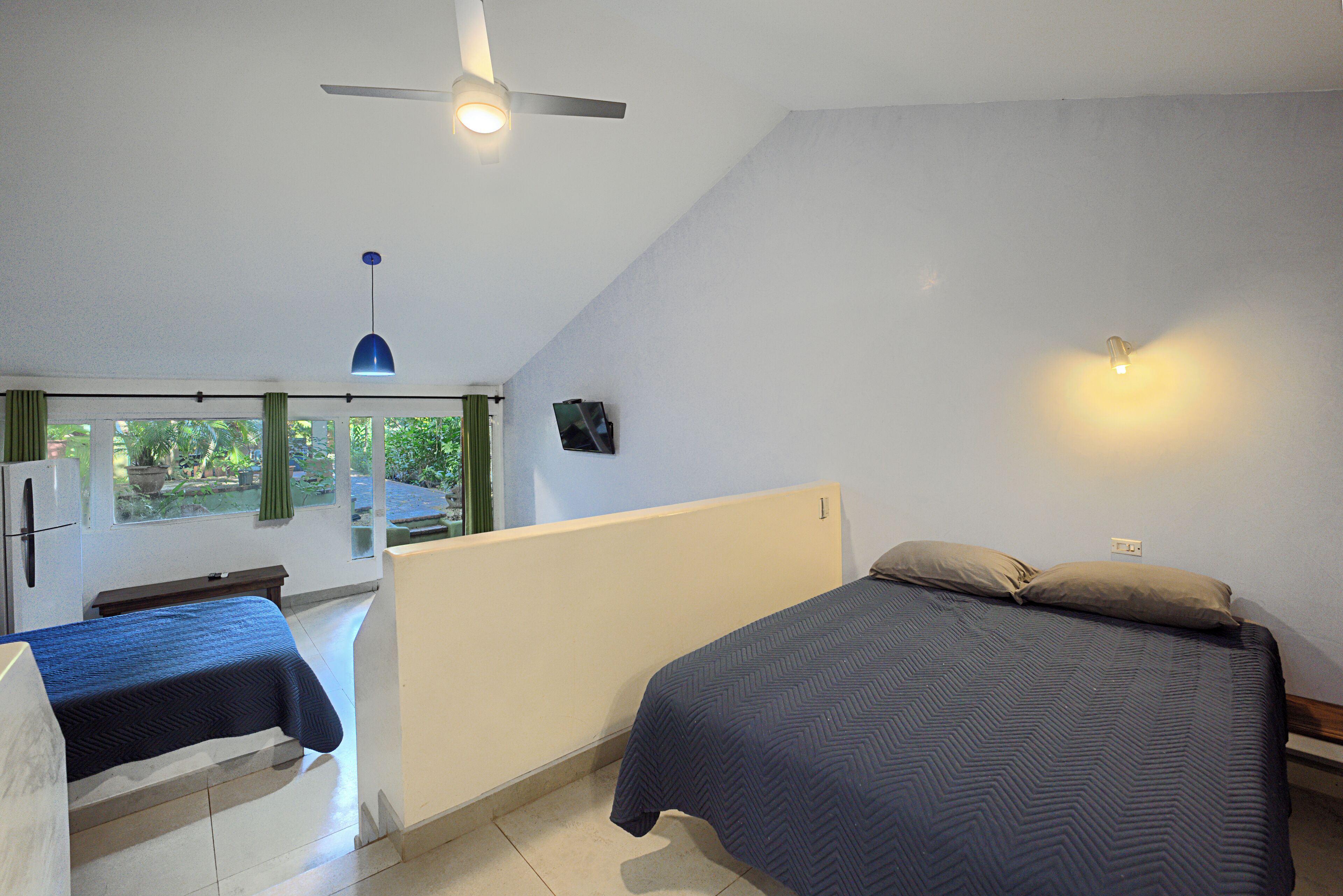 Une chambre à coucher avec un lit, un bureau et une chaise. Il y a une fenêtre avec des rideaux, un ventilateur de plafond et un luminaire fixé au mur.