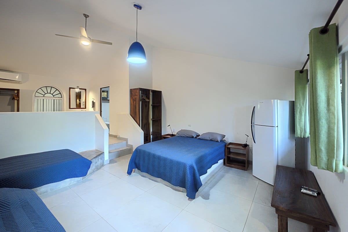 Un dormitorio con dos camas, ventilador de techo, mesita de noche, espejo, ventana con cortinas y un refrigerador.