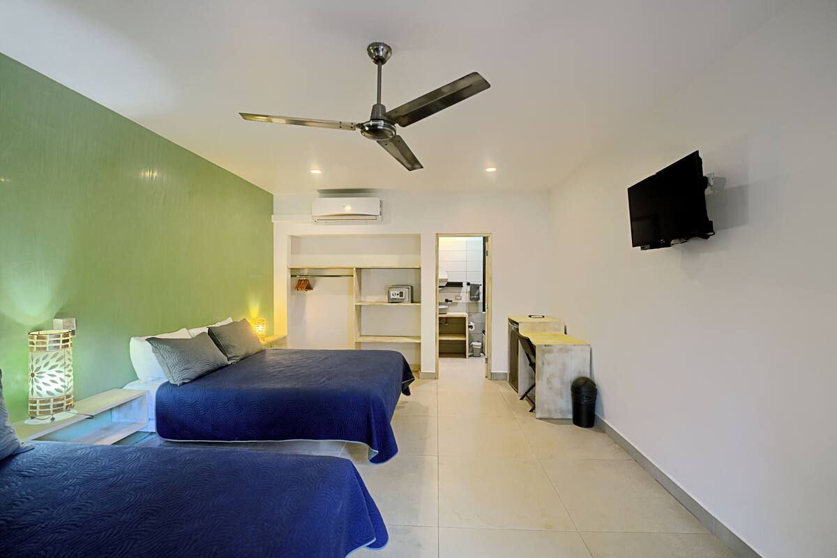 Habitación de hotel con dos camas, ventilador de techo, televisión montada en la pared y una pequeña zona de kitchenette.