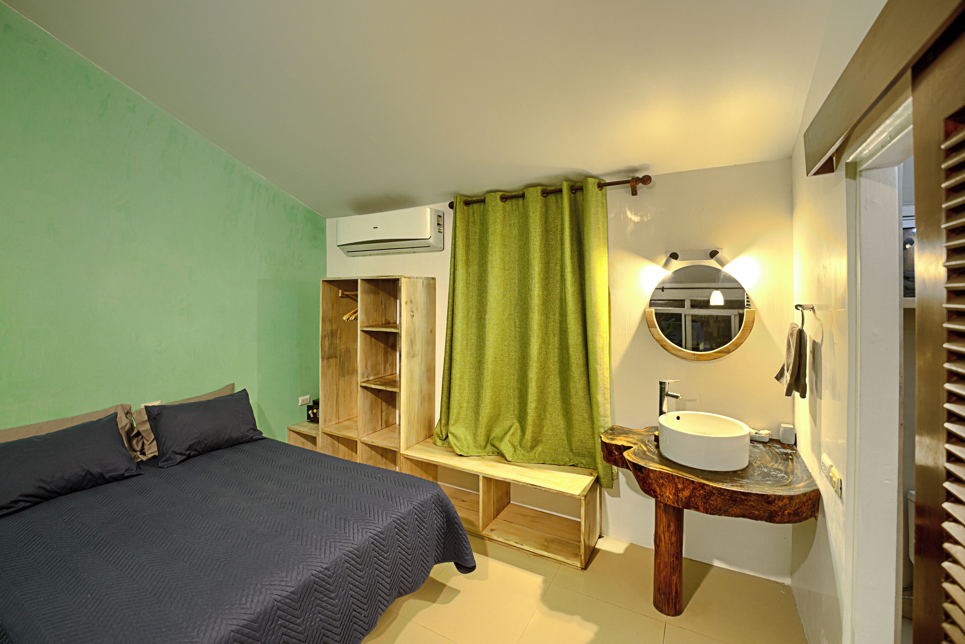A bedroom with a bed, a wooden bench, a green curtain, a round mirror, and a sink with a wooden countertop.