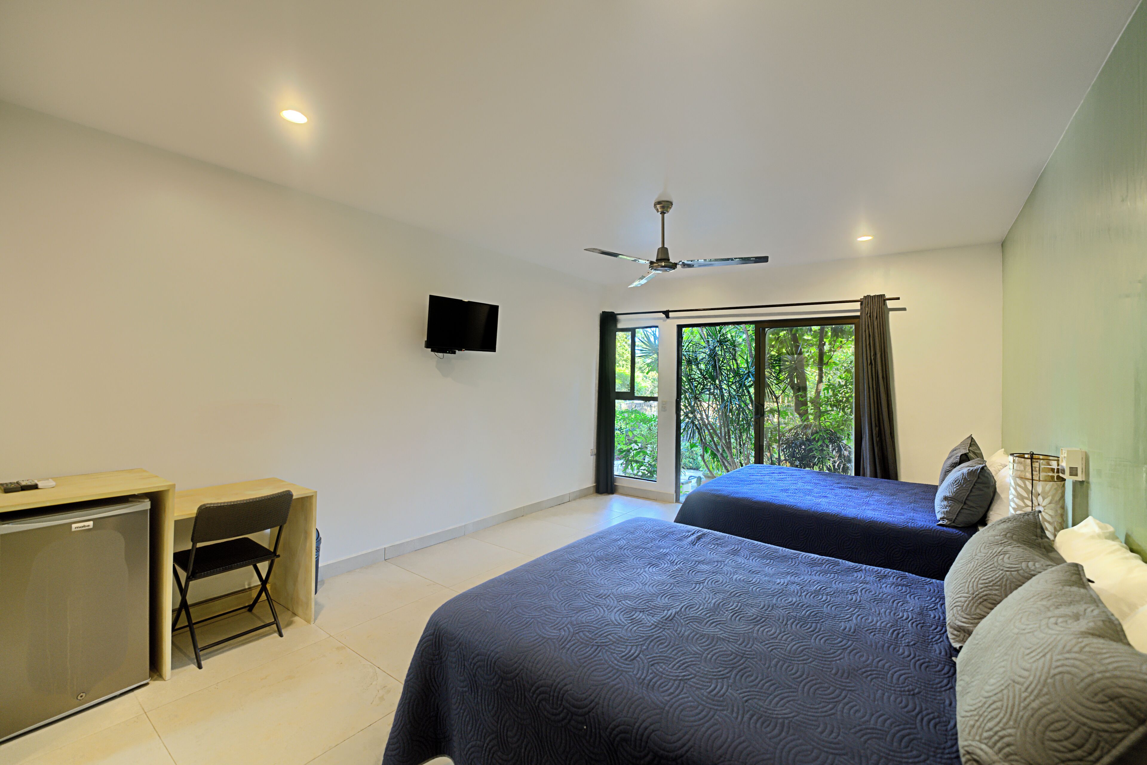 A hotel room with two beds, a desk, a chair, a television, and a ceiling fan.