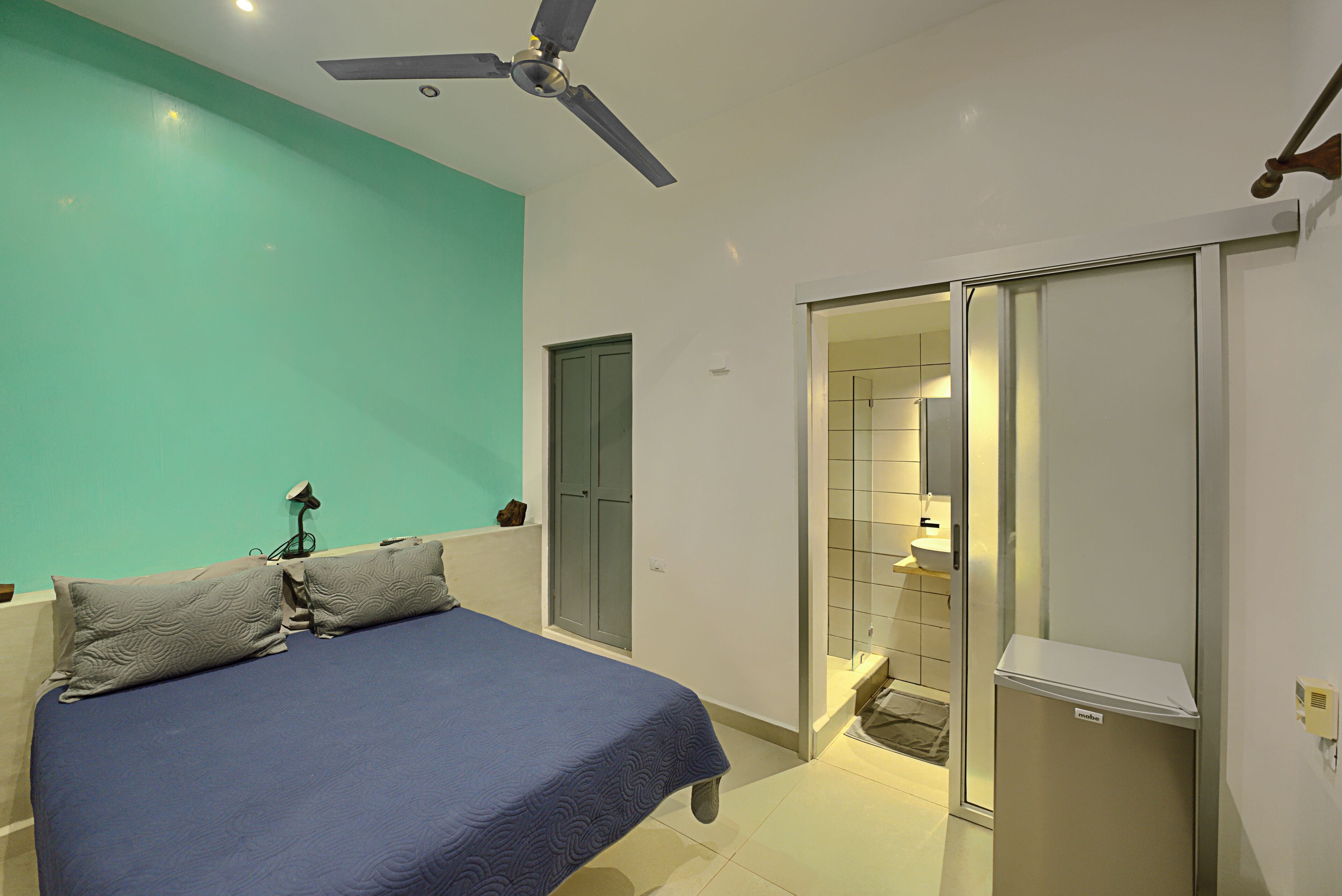 Un dormitorio con cama, ventilador de techo, armario y baño visible a través de una puerta de cristal.