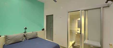 Un dormitorio con cama, ventilador de techo, armario y baño visible a través de una puerta de cristal.