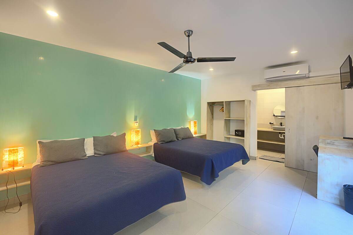 Habitación de hotel con dos camas, ventilador de techo, unidad de aire acondicionado y baño visible a través de una puerta abierta.